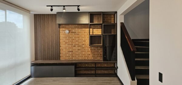 Cada espacio es único, y por ello diseñamos muebles personalizados que se adaptan tanto a la arquitectura como a las necesidades del usuario. Este ejemplo integra materiales como madera, ladrillo expuesto y elementos metálicos para crear una atmósfera cálida y contemporánea. La composición modular aprovecha al máximo el muro disponible y aporta funcionalidad con áreas de almacenamiento abiertas y cerradas, ideal para espacios habitacionales o de trabajo.<br />
<a href="/cajica" class="btn-proyecto" target="_blank">Ver Proyecto</a> Diseño y fabricación de mobiliario a medida