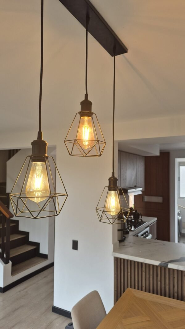 Una iluminación colgante de estilo industrial moderno destaca en este ambiente cálido y acogedor. Las lámparas de filamento, con estructuras geométricas metálicas, aportan carácter y personalidad al espacio, creando un contraste elegante con los tonos neutros de la cocina y el comedor. La combinación de madera, hierro y luz cálida genera una atmósfera contemporánea con un toque artesanal, ideal para disfrutar de un hogar funcional y estéticamente equilibrado.<br />
<a href="/cajica" class="btn-proyecto" target="_blank">Ver Proyecto</a> Diseño y ambiente: