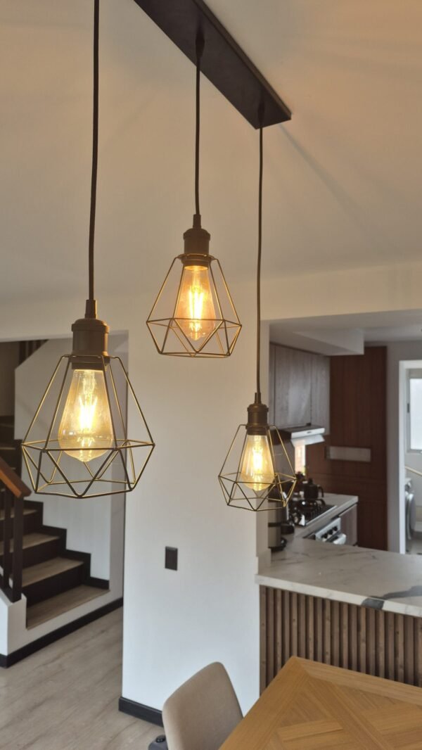 Una iluminación colgante de estilo industrial moderno destaca en este ambiente cálido y acogedor. Las lámparas de filamento, con estructuras geométricas metálicas, aportan carácter y personalidad al espacio, creando un contraste elegante con los tonos neutros de la cocina y el comedor. La combinación de madera, hierro y luz cálida genera una atmósfera contemporánea con un toque artesanal, ideal para disfrutar de un hogar funcional y estéticamente equilibrado.<br />
<a href="/cajica" class="btn-proyecto" target="_blank">Ver Proyecto</a> Diseño y ambiente: