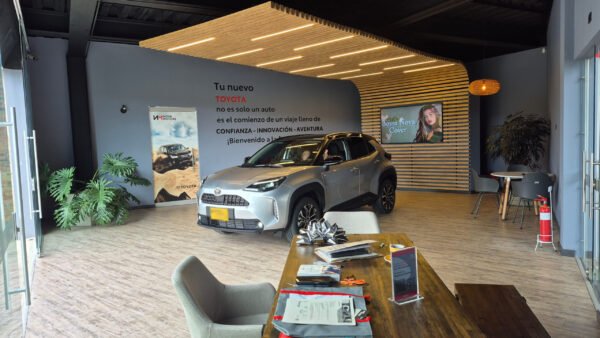 El diseño del showroom destaca por una paleta de grises neutros contrastada con el acabado cálido tipo madera del piso y la estructura curva del fondo. Esta última, hecha en listones de madera con iluminación cálida integrada, crea un punto focal elegante y natural. Las lámparas colgantes tejidas refuerzan el estilo acogedor y artesanal. La vegetación interior y la luz natural completan un ambiente moderno y atractivo.<a href="/toyota" class="btn-proyecto" target="_blank">Ver Proyecto</a> SALA DE ENTREGA